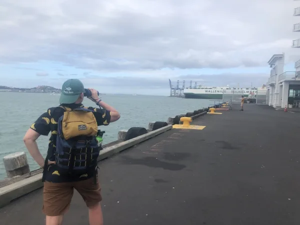 Gillian Murphey conducting part of the Human Uses of Ocean Space Census, looking over at container ship cranes and an auto carrier in Auckland, Aotearoa New Zealand.